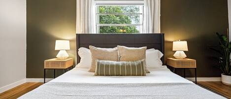 Une chambre avec un grand lit, deux tables de chevet avec des lampes, une fenêtre avec des rideaux et une vue sur la verdure à l’extérieur.