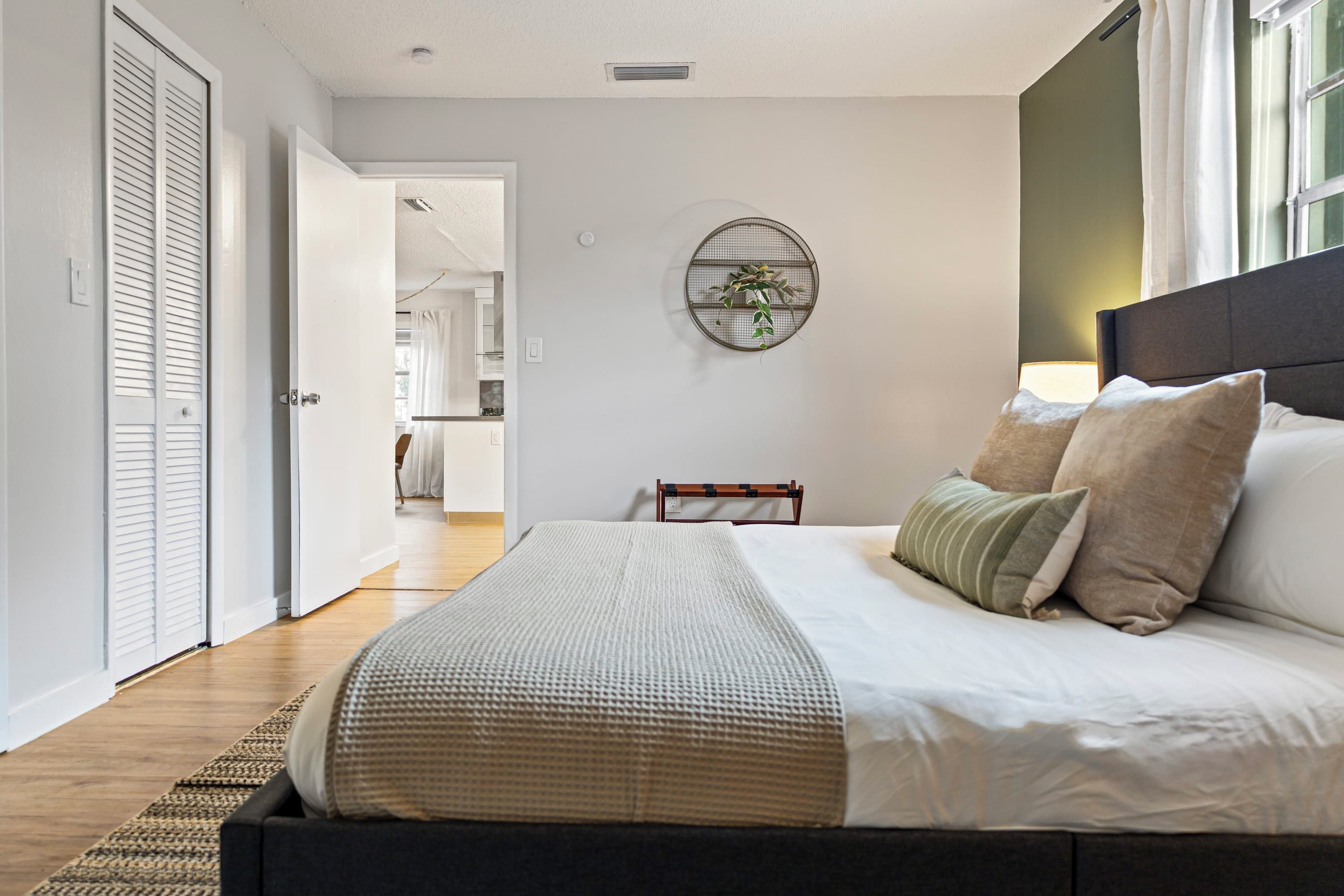 Quarto com cama, travesseiros, mesa de cabeceira, planta na parede e vista para um corredor.