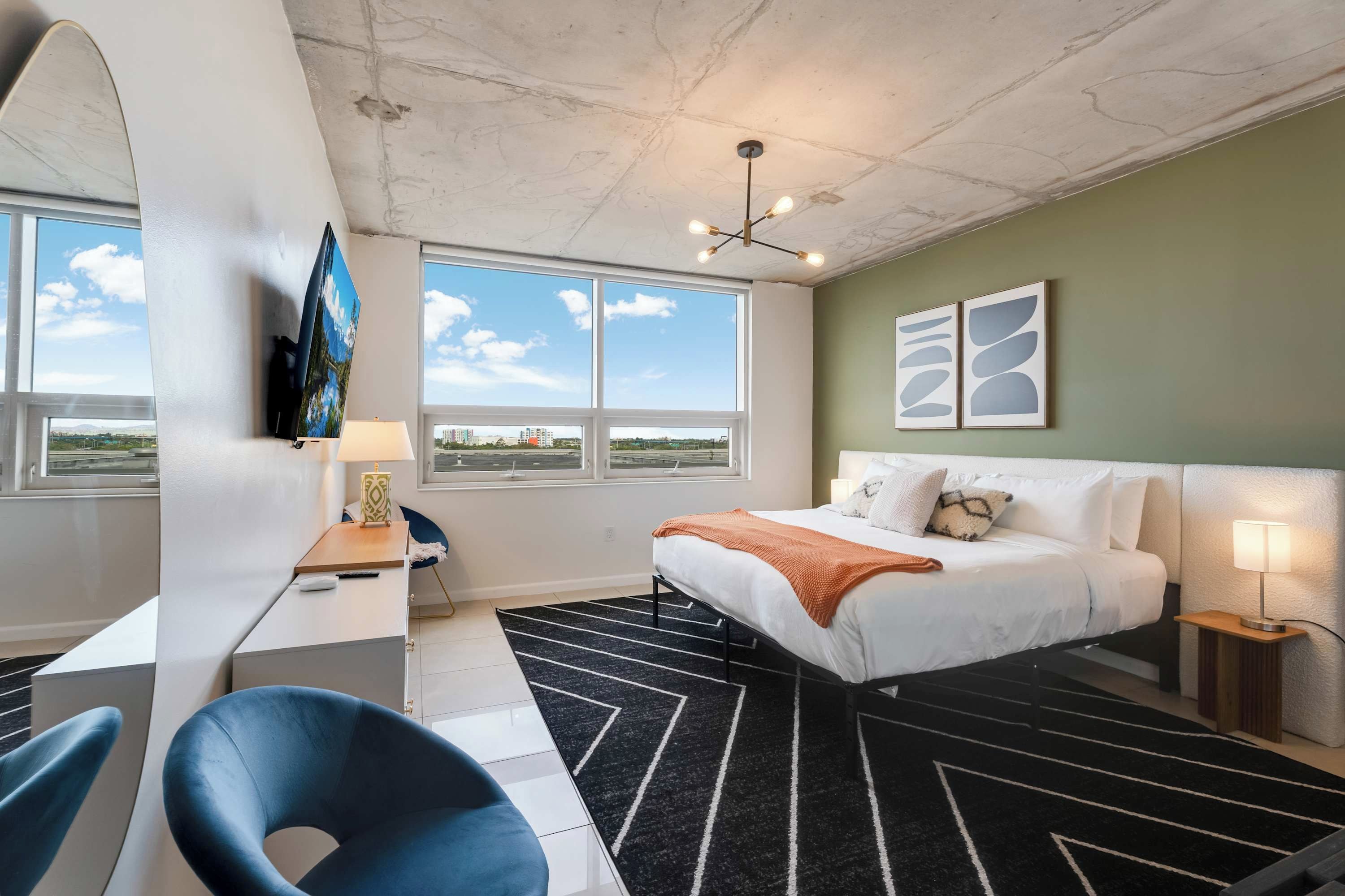 Quarto de hotel com cama, escrivaninha, cadeira e janelas amplas.