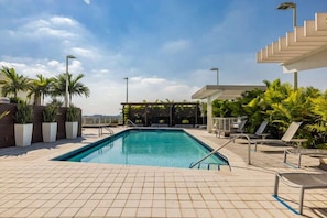 Una piscina en la azotea con suelo de baldosas, sillas de descanso y palmeras.