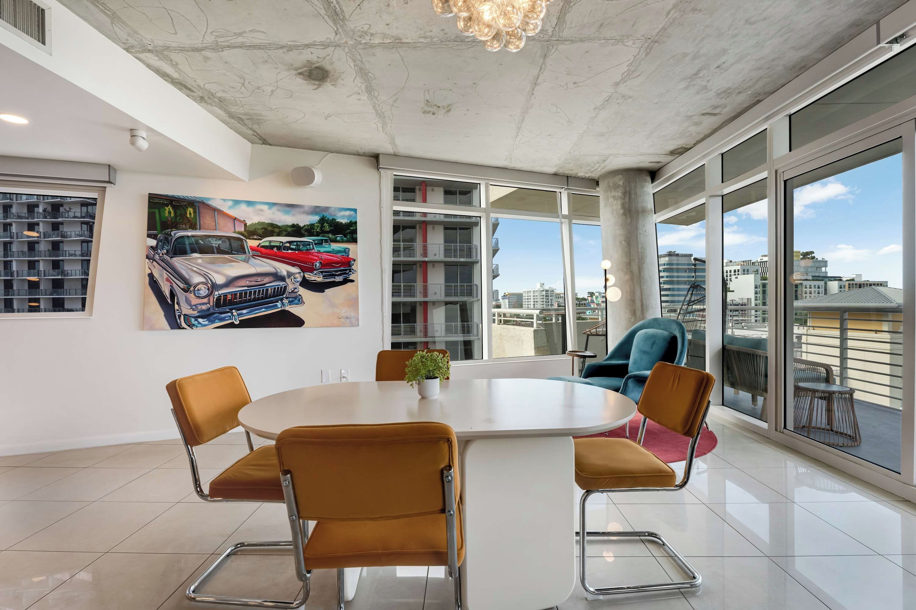 Uma área de jantar moderna com uma mesa redonda, quatro cadeiras, uma planta em vaso e vista para a cidade através de grandes janelas.