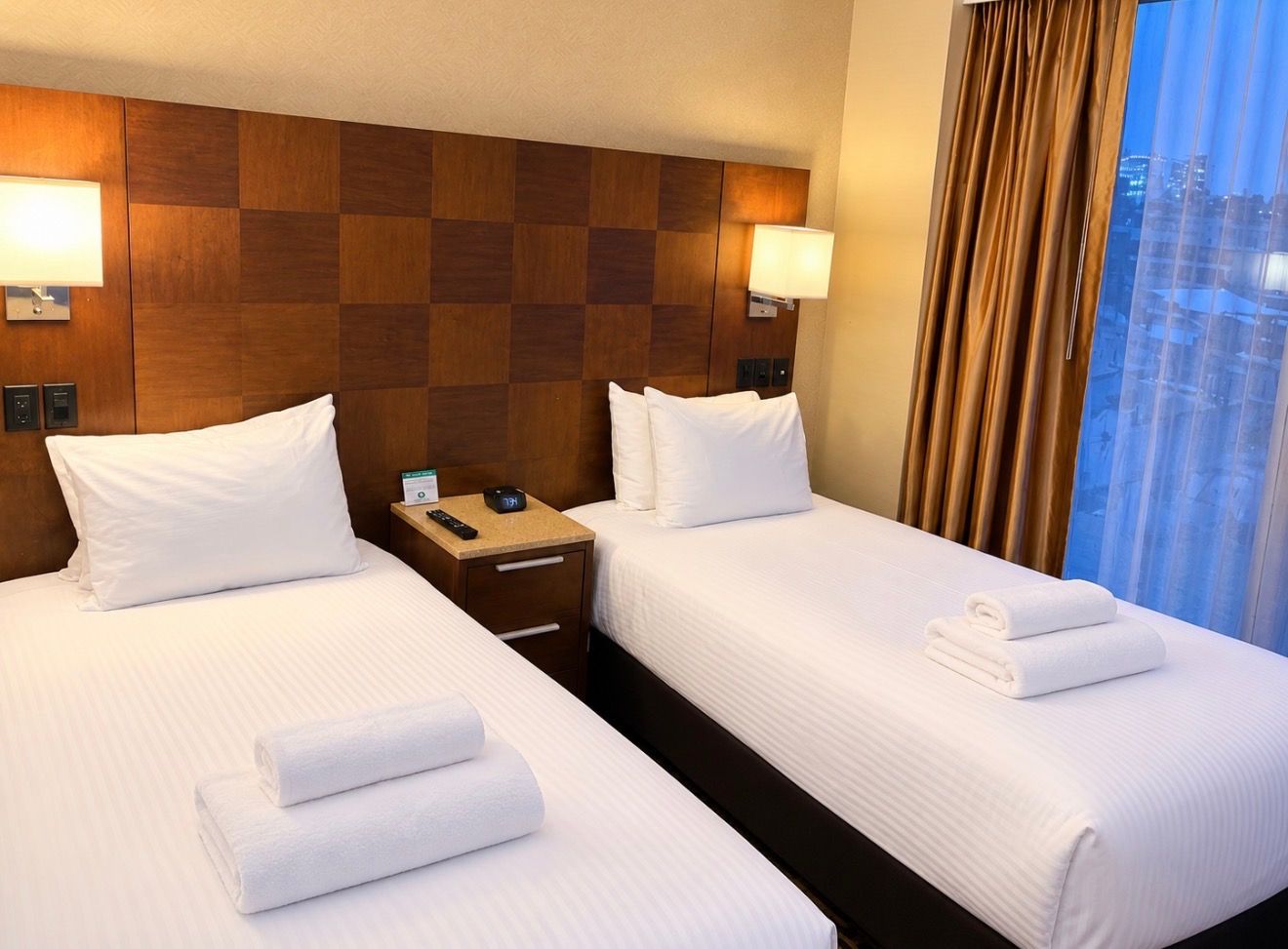 A hotel room with two beds, each with white linens and pillows, a wooden headboard, bedside lamps, a nightstand with a remote control, and a window with curtains.