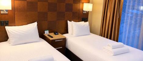 A hotel room with two beds, each with white linens and pillows, a wooden headboard, bedside lamps, a nightstand with a remote control, and a window with curtains.