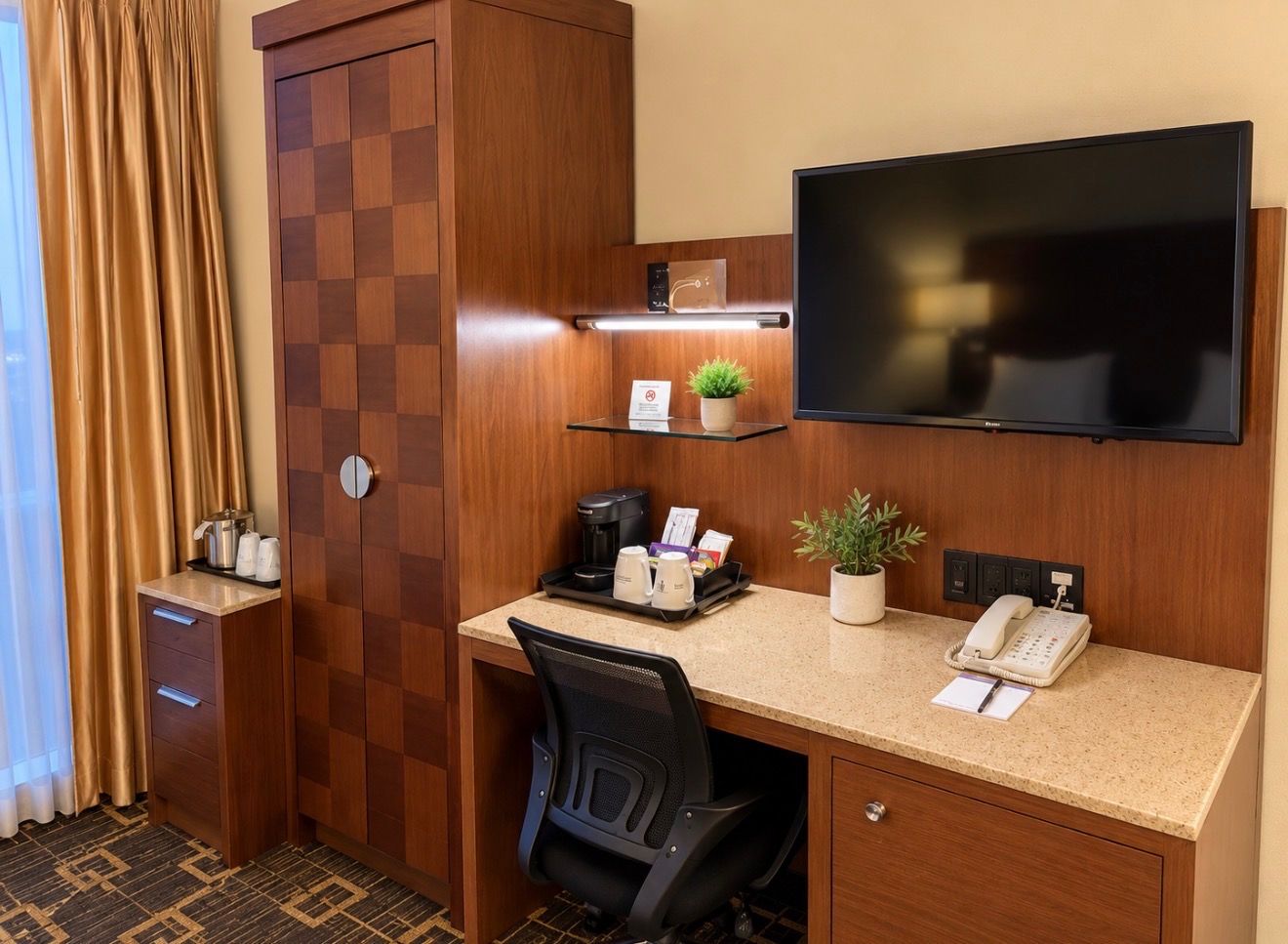 Habitación de hotel con escritorio, televisor de pantalla plana, teléfono, cafetera, planta y minibar.