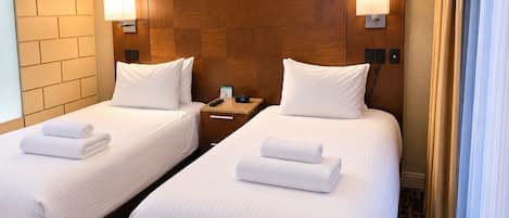 Habitación de hotel con dos camas, cada una con ropa blanca y toallas, cabeceros de madera, lámparas de noche y mesitas de noche con teléfono y reloj.