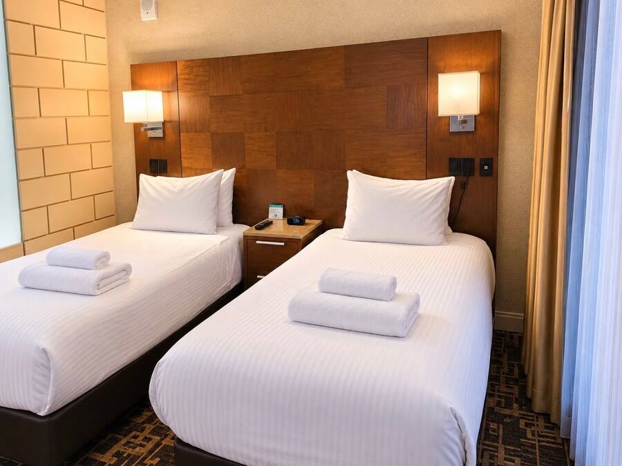 Quarto de hotel com duas camas, cada uma com lençóis e toalhas brancos, cabeceira de madeira, luminárias laterais e mesa de cabeceira com telefone e relógio.