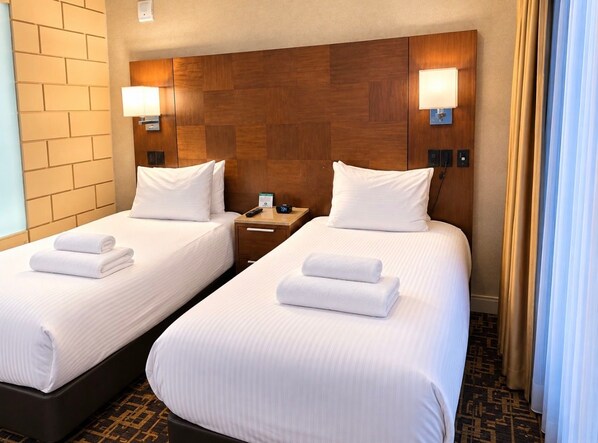 A hotel room with two beds, each with white linens and towels, a wooden headboard, bedside lamps, and a small bedside table with a phone and a clock.