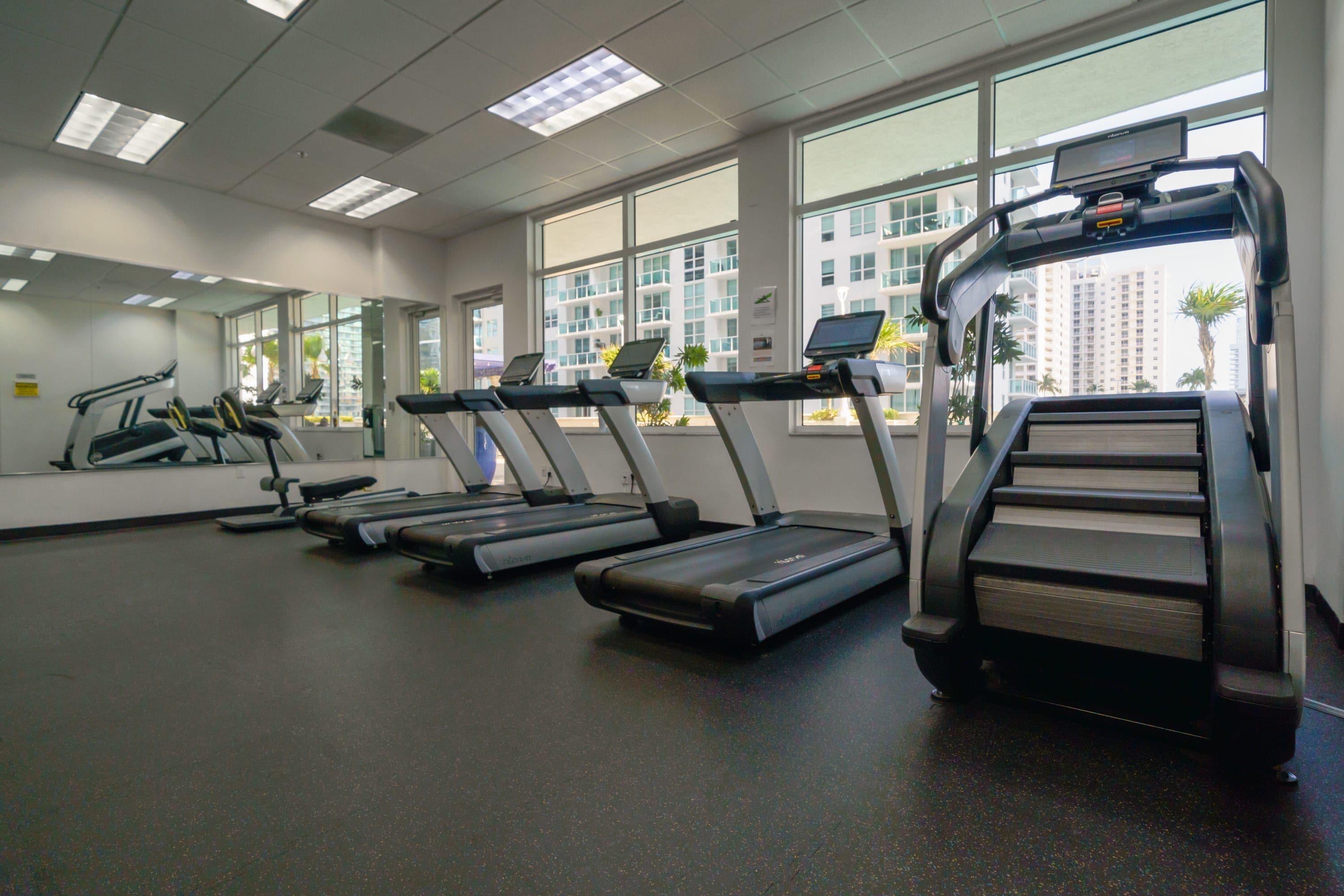 A gym with treadmills, a mirror, and large windows with a city view.