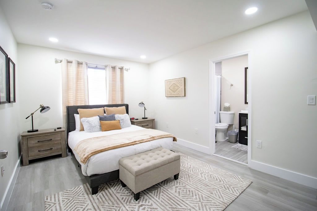 Quarto com cama, mesas de cabeceira, um banquinho, janela com cortinas, um quadro na parede e um banheiro visível por uma porta aberta.