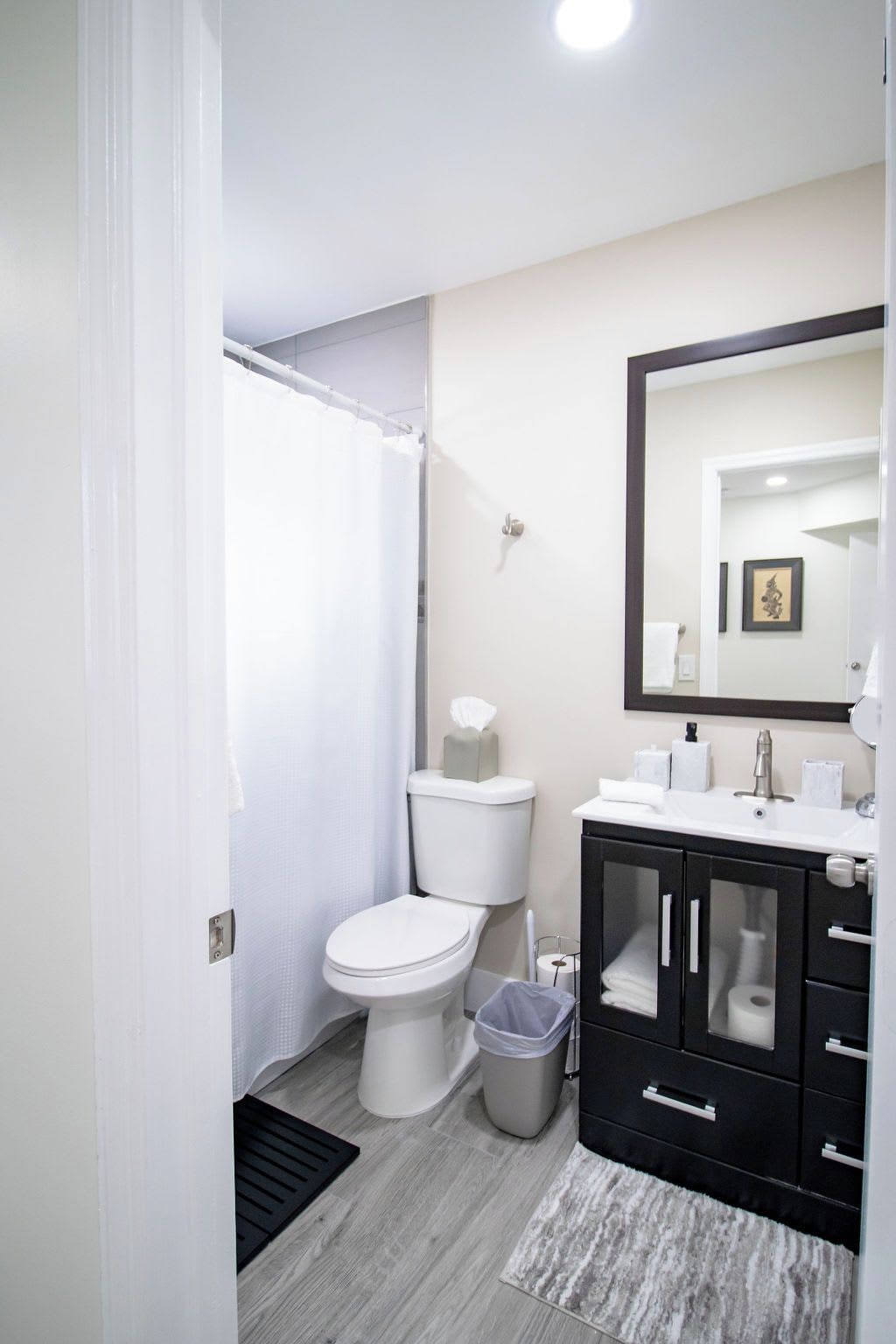 Un baño con inodoro, cortina de ducha, lavamanos con espejo y un cubo de basura.