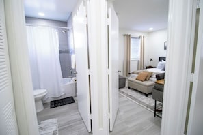 Uma casa de banho moderna com sanita, chuveiro e uma porta branca que dá para um quarto com cama e secretária.