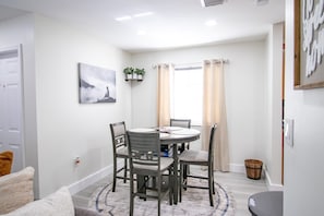 A dining area with a round table, four chairs, a small plant shelf, a framed picture, and a window with curtains.