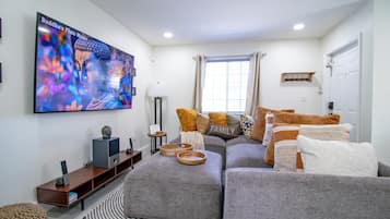 Una sala de estar moderna con un televisor de pantalla plana grande, sofás grises, una alfombra estampada y una ventana con cortinas.