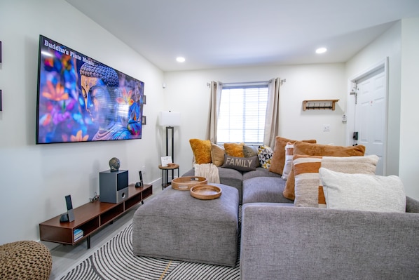 A modern living room with a large flat-screen TV, grey sofas, a patterned rug, and a window with curtains.