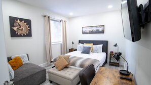 A hotel room with a bed, a desk, a chair, a small ottoman, a nightstand, a lamp, a television, and artwork on the walls. - Good Vibes Oasis, 2 Story Townhome in Cutler Bay (Cutler Bay)