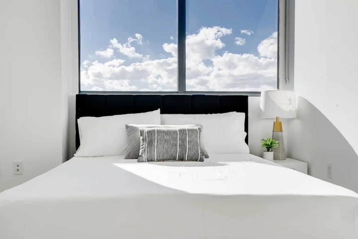 A bed with white bedding, a striped pillow, a nightstand with a lamp, and a small plant.