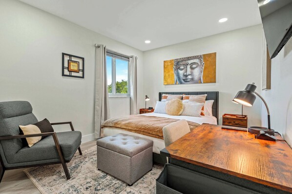A bedroom with a bed, a chair, a desk, a lamp, a window with curtains, a painting on the wall, and a small table.