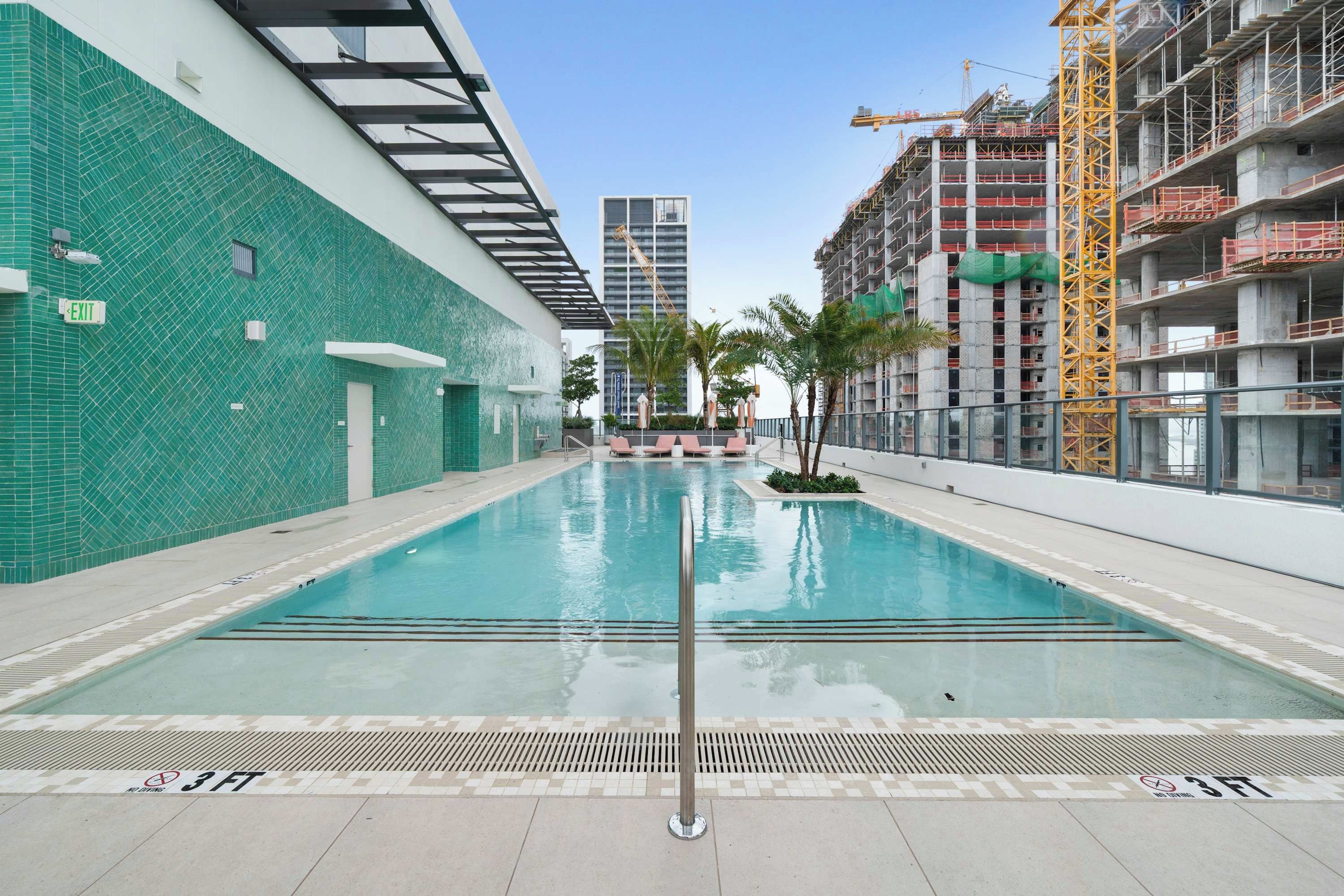 A swimming pool with a 3 FT depth marker, surrounded by a building with a green tiled exterior and a construction crane in the background.