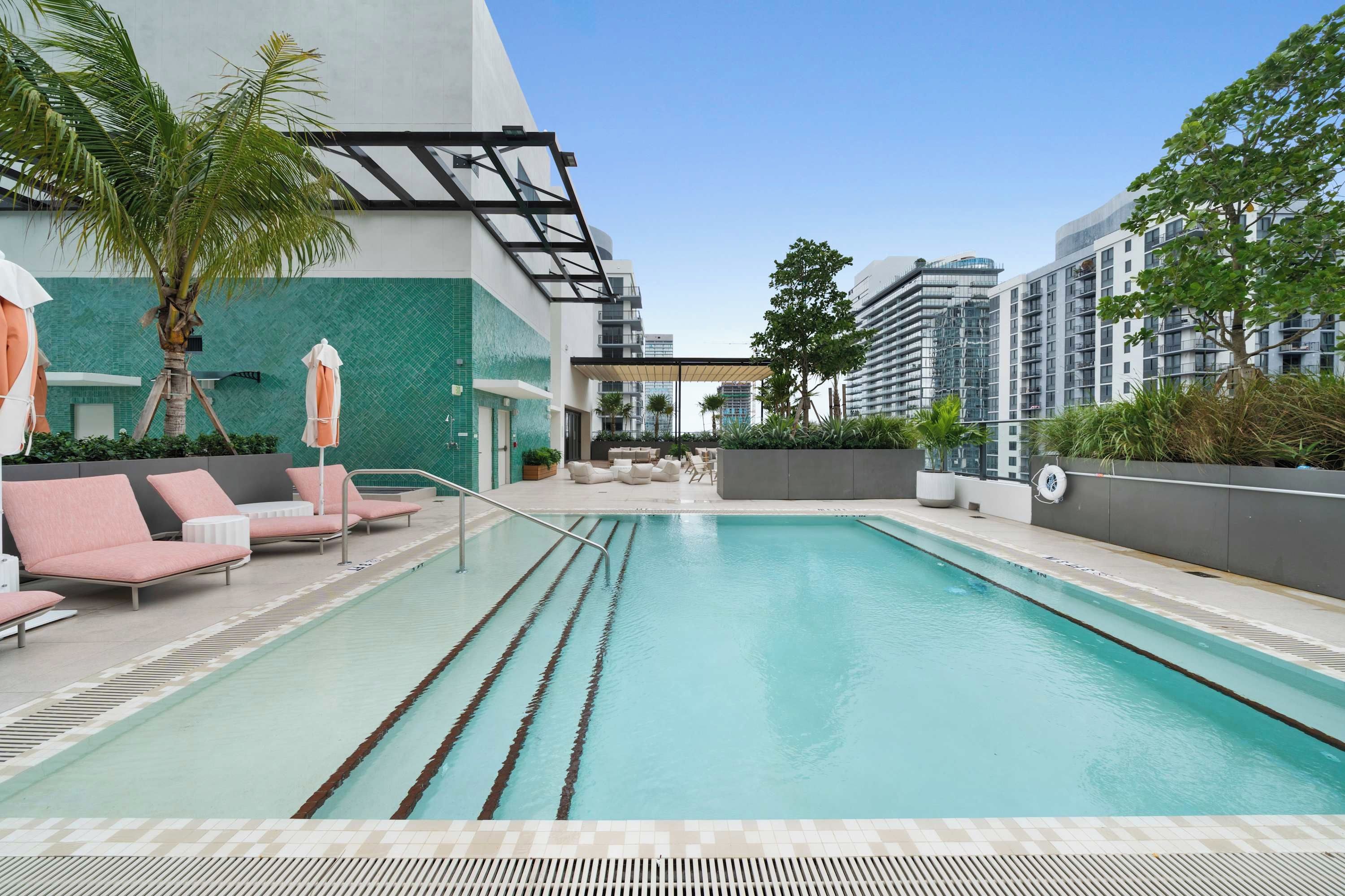 A swimming pool with lounge chairs and umbrellas, surrounded by greenery and modern buildings.