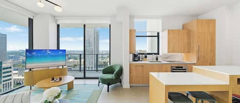 A modern apartment with a living area featuring a TV, sofa, and coffee table, a kitchen with wooden cabinets, and large windows offering a city view.