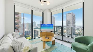 A modern living room with a large TV, a sofa, a green armchair, and a view of a cityscape through large windows.