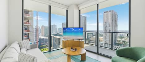 A modern living room with a large TV, a sofa, a green armchair, and a view of a cityscape through large windows.