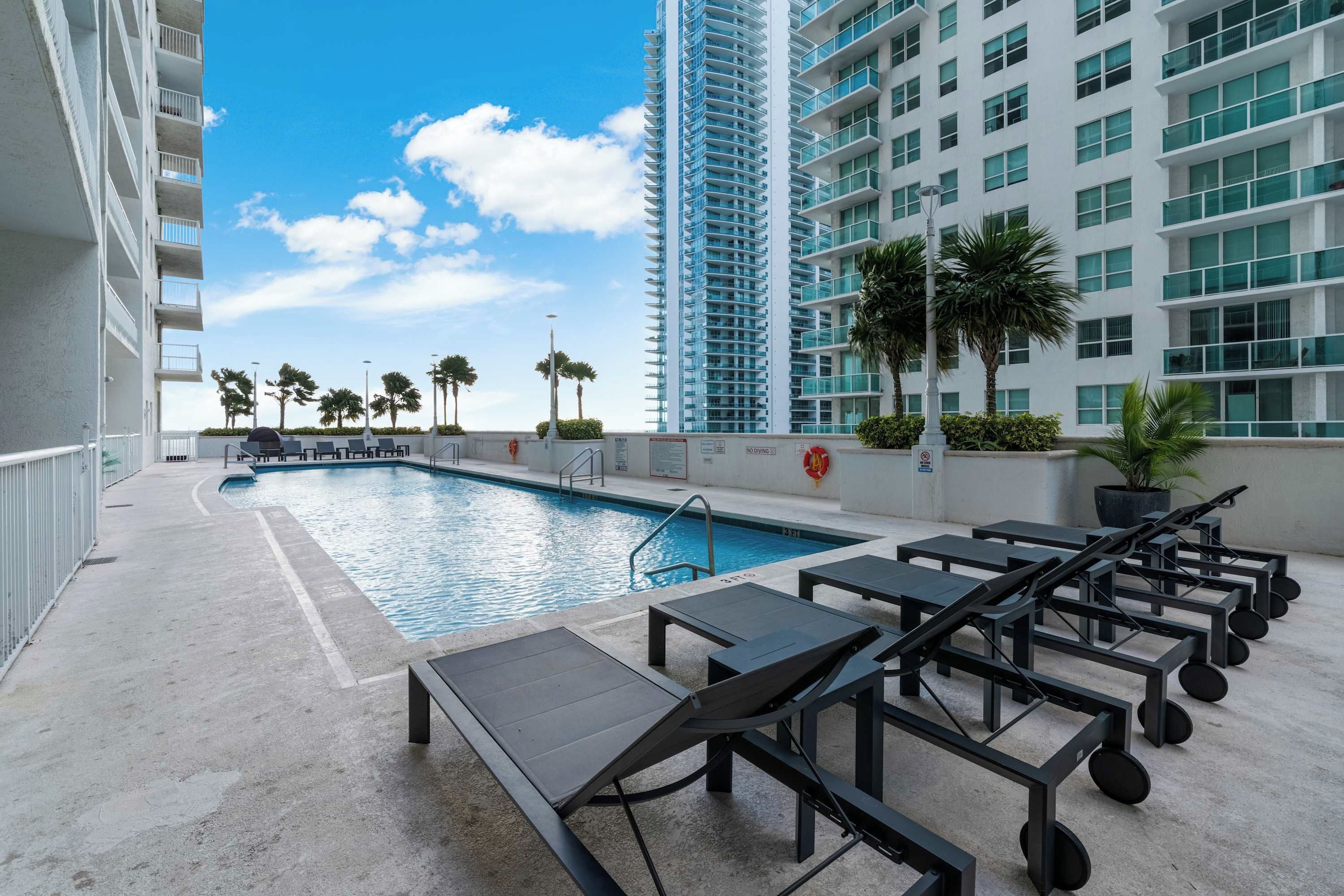 A rooftop pool area with sun loungers and a swimming pool, surrounded by high-rise buildings and palm trees.
