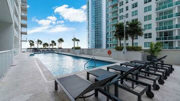 A rooftop pool area with sun loungers and a swimming pool, surrounded by high-rise buildings and palm trees.