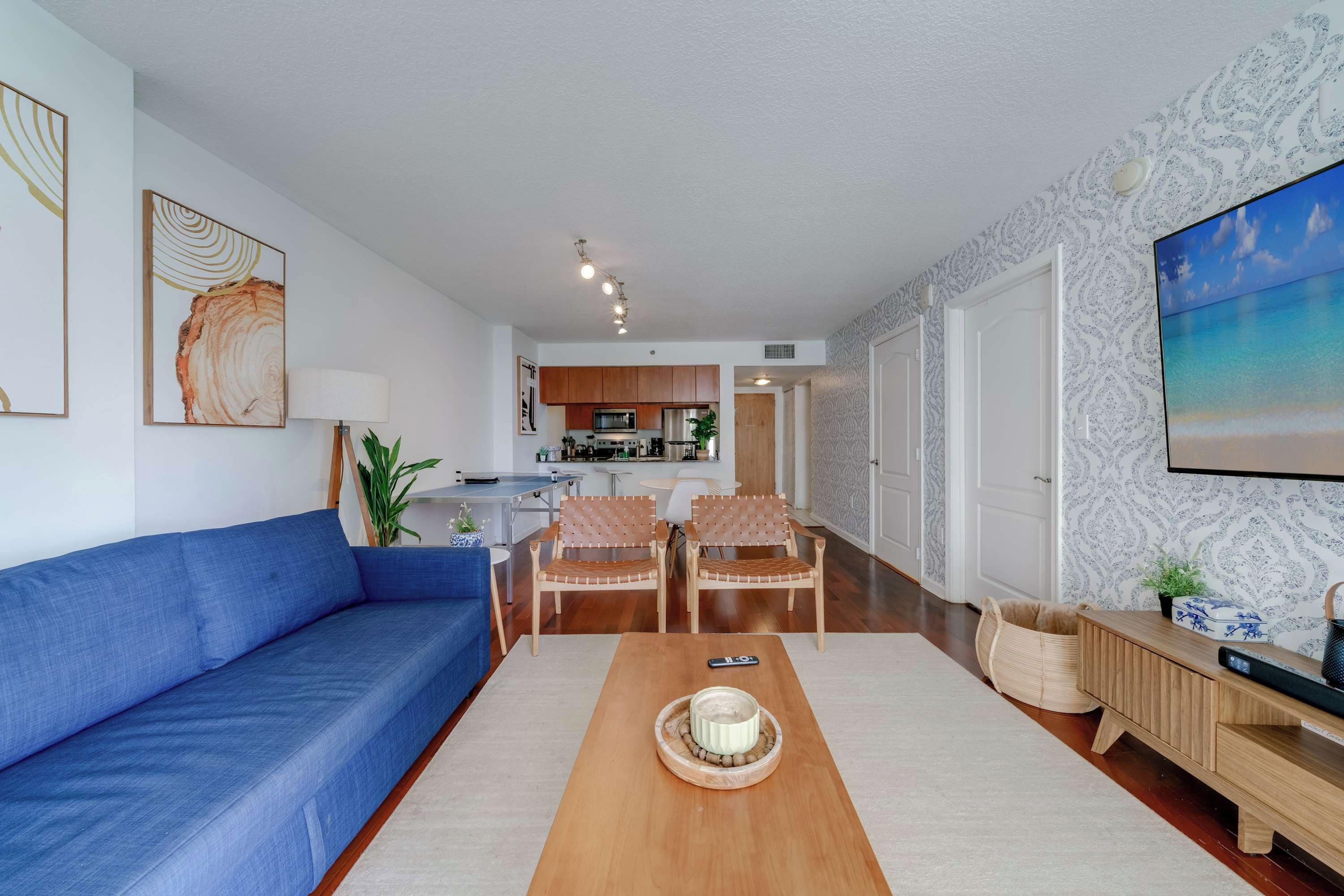 A modern living room with a blue sofa, wooden coffee table, and wicker chairs. A kitchen is visible in the background with wooden cabinets and a microwave.