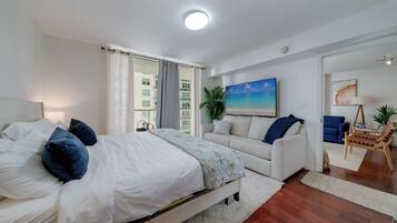 A bedroom with a bed, a sofa, a chair, a painting, and a plant.