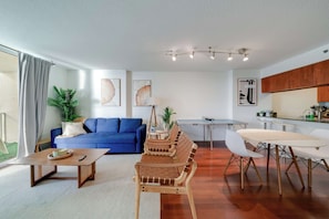 Uma sala de estar moderna com um sofá azul, mesa de centro de madeira e uma área de jantar com mesa e cadeiras brancas.