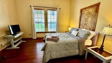 Un dormitorio con cama, televisión sobre un mueble bajo, ventana con cortinas y un cuadro en la pared.