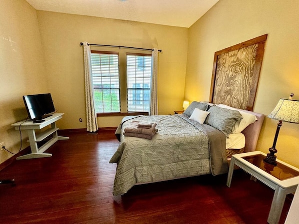 A bedroom with a bed, a television on a stand, a window with curtains, and a painting on the wall.