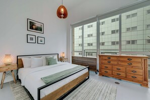 A modern bedroom with a large bed, wooden dresser, and a view of a building through large windows. - The Vizcayne Vibe Sauna, Pool and Sundeck (Miami)