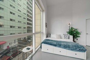 Ein modernes Schlafzimmer mit einem großen Fenster, einem Bett mit blauer Decke, einem Blumentopf und einem kleinen Tisch mit einem Stuhl.