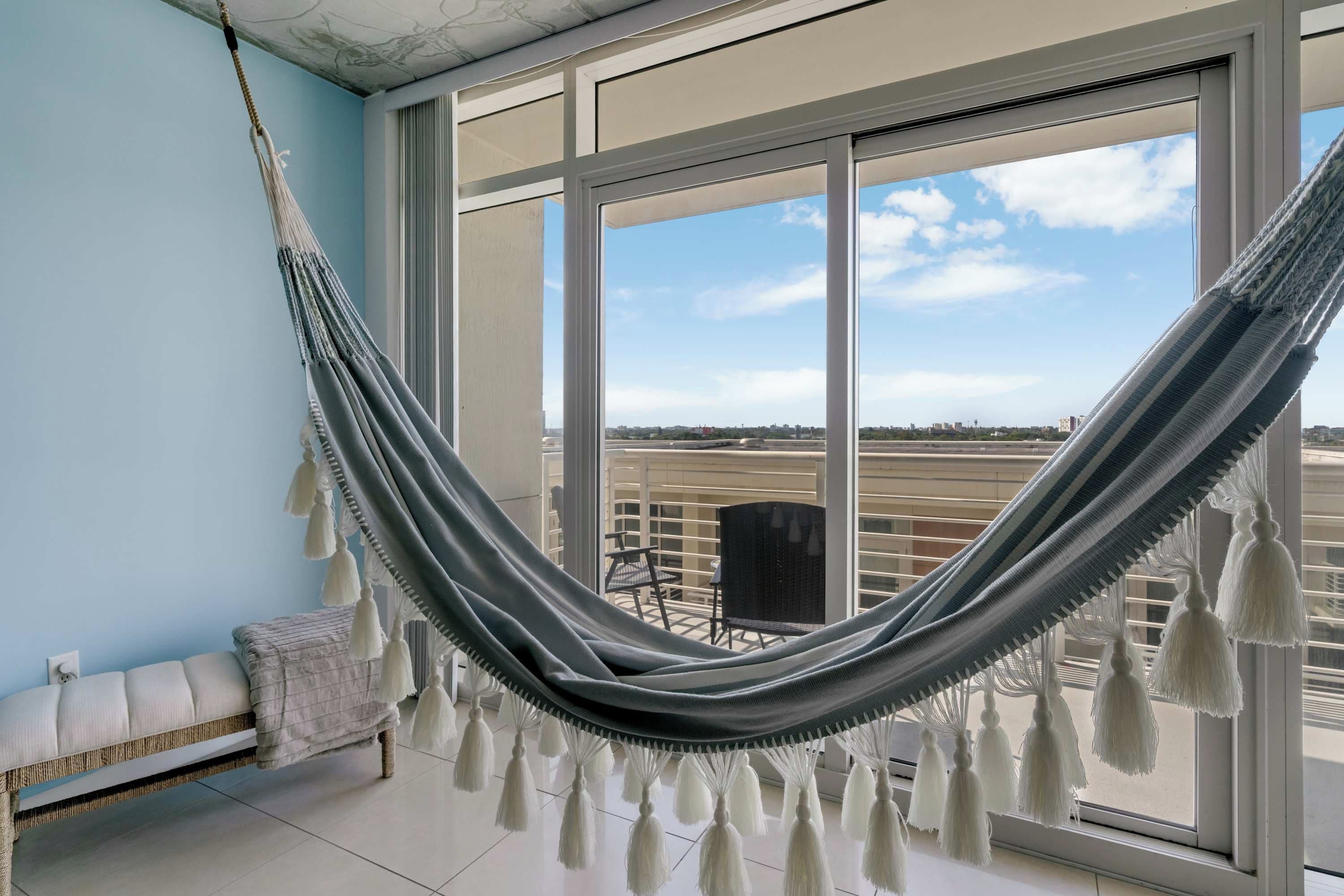 A hammock in a room with a large window, a bench, and a view of the city.