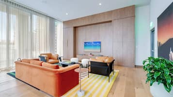 A modern living room with a large orange sofa, a black armchair, and a small table. There is a large window with sheer curtains, a painting on the wall, and a potted plant.