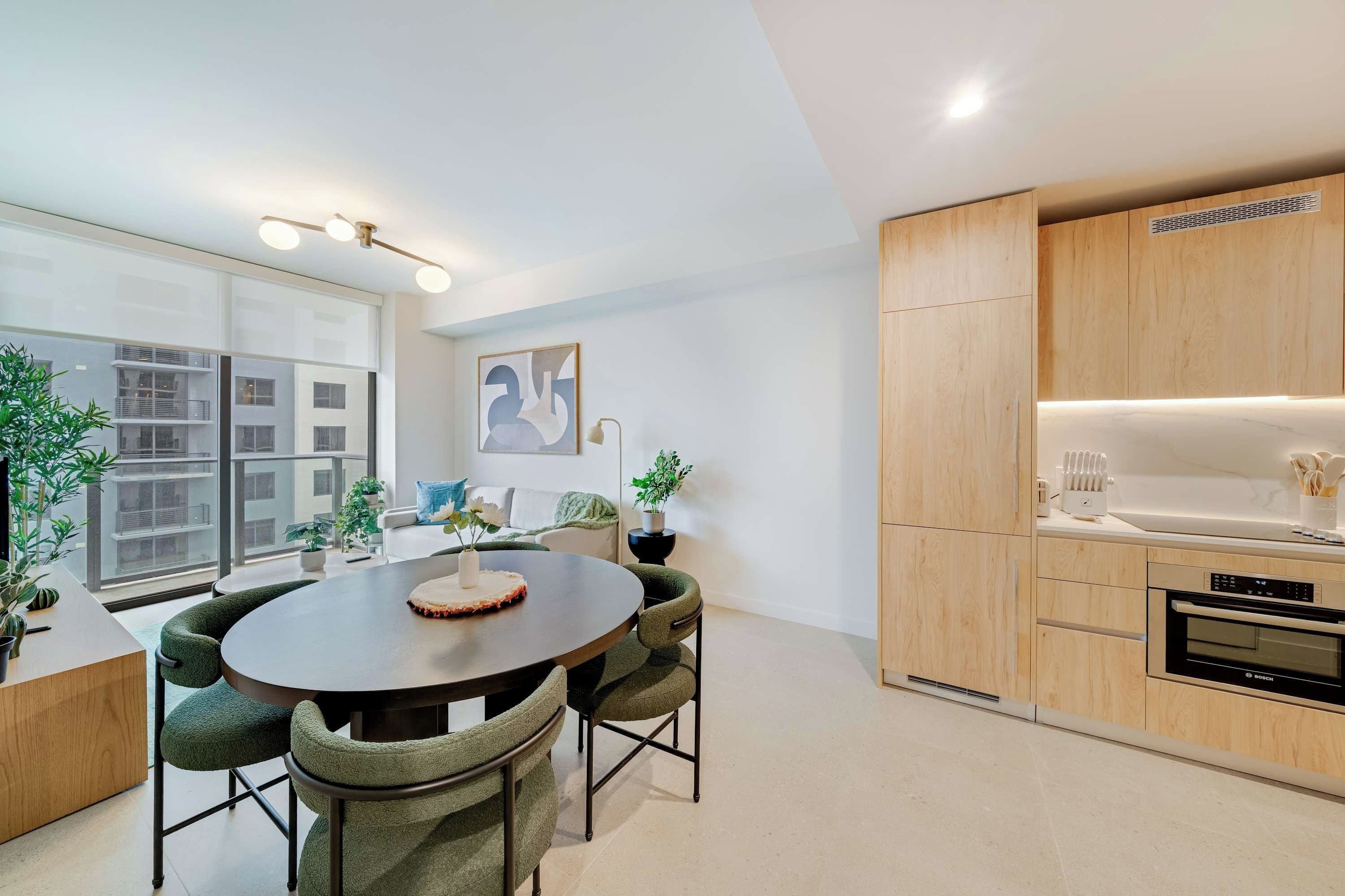 A modern kitchen with wooden cabinets, a built-in oven, and a dining area with a round table and green upholstered chairs.