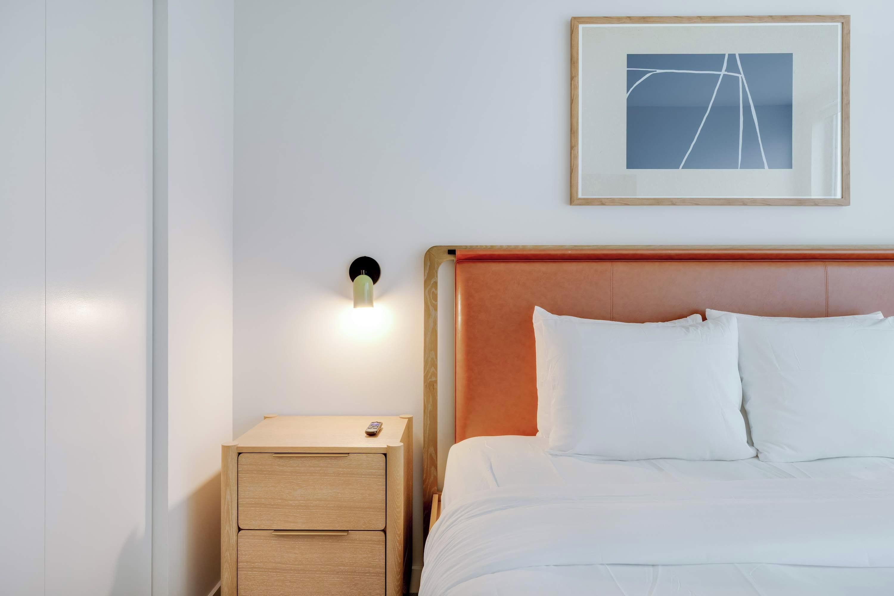 A neatly made bed with white linens, a wooden nightstand with drawers, a framed picture on the wall, and a wall-mounted light.