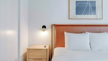 A neatly made bed with white linens, a wooden nightstand with drawers, a framed picture on the wall, and a wall-mounted light.