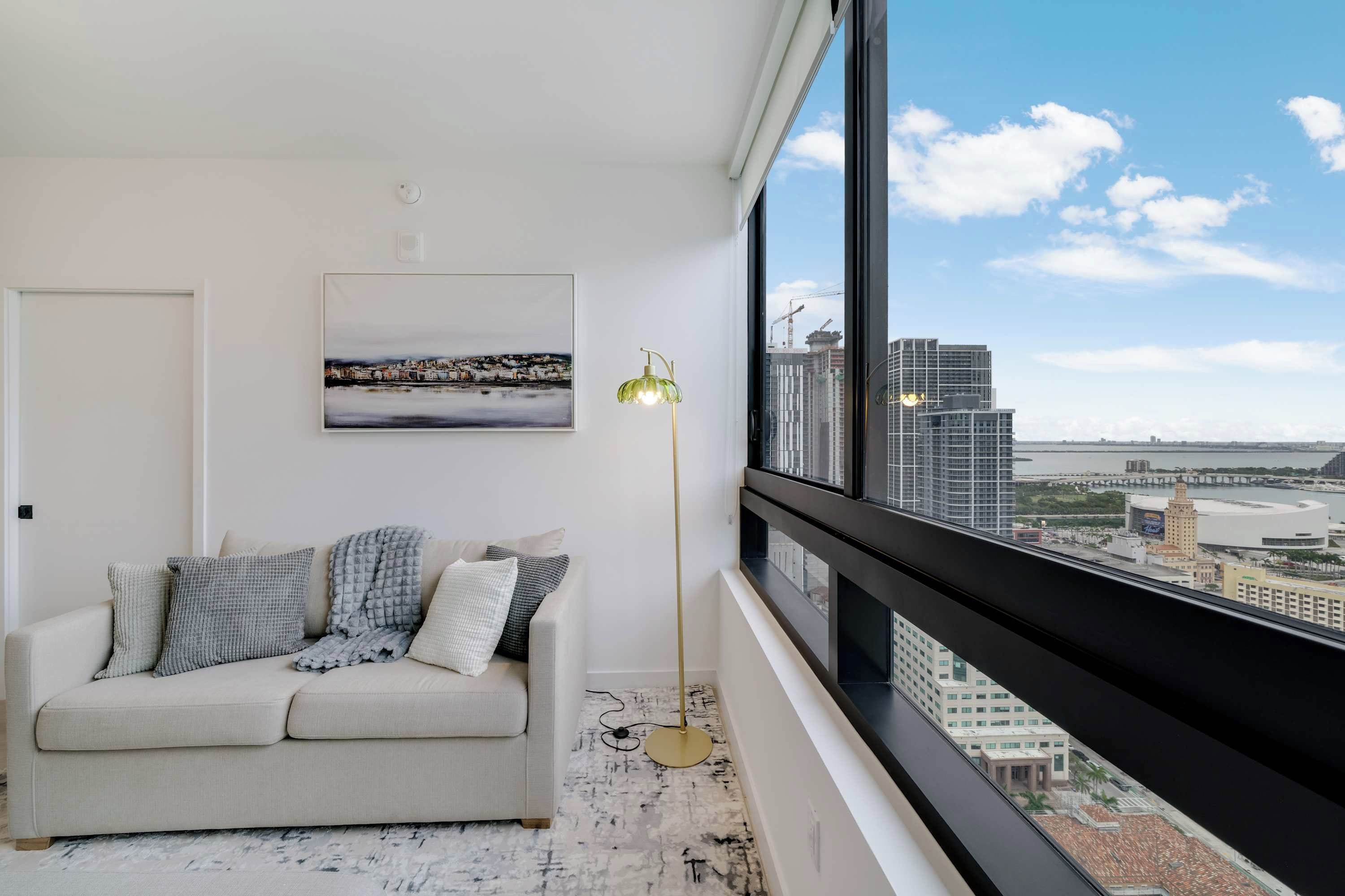 A modern living room with a beige sofa, a floor lamp, and a cityscape view through large windows.