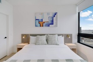 A modern hotel room with a large bed, two bedside tables, a headboard with integrated lighting, a window with a city view, and a geometric abstract painting on the wall. - City Atelier Downtown, Sauna, Pool, Parking (Miami)
