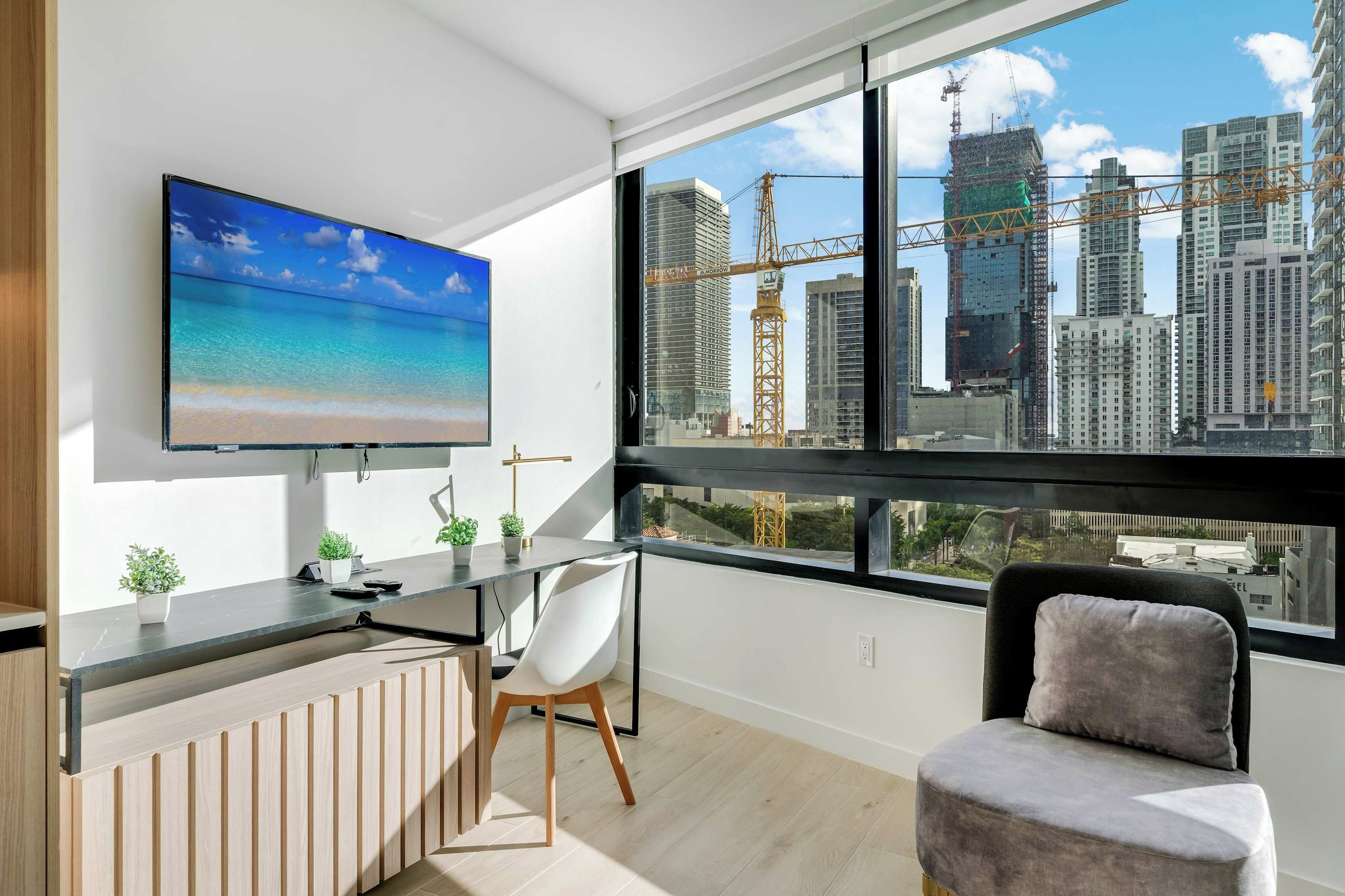 A modern room with a large TV, a desk, a chair, and a view of a cityscape with buildings and a crane.