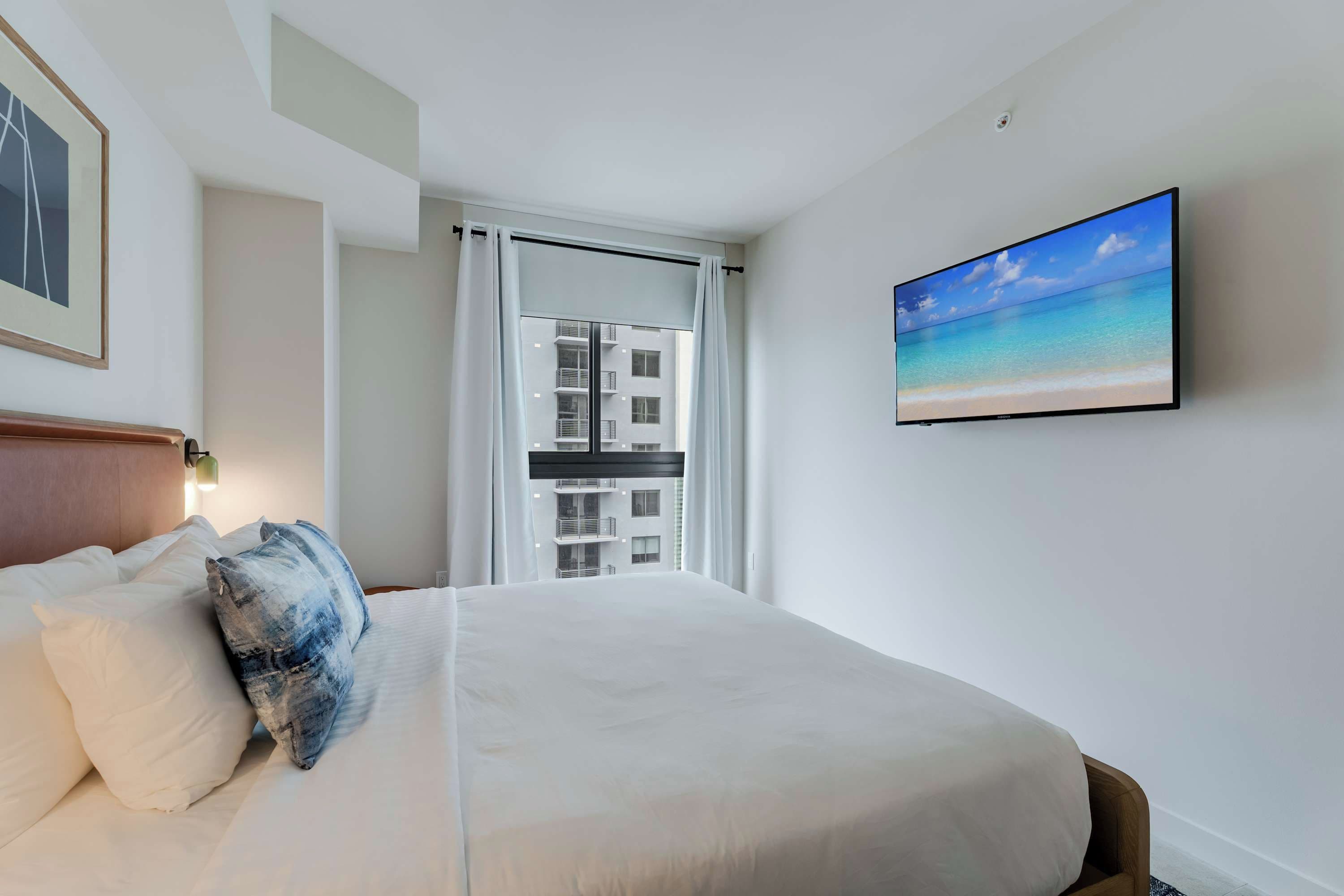 A hotel room with a bed, a painting of a beach, and a view of a building through a window.