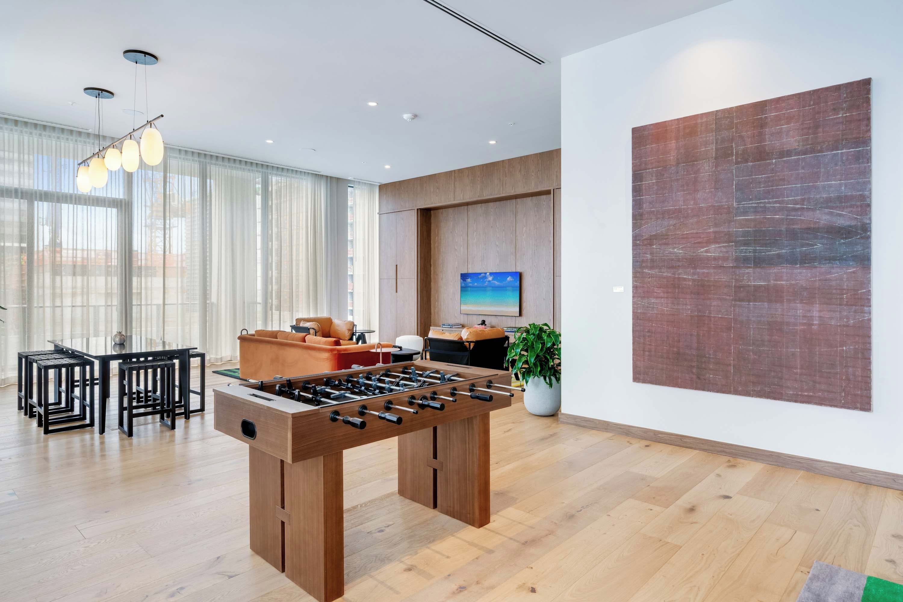 A modern room with a wooden foosball table, a dining area with black chairs, and a large abstract painting on the wall.