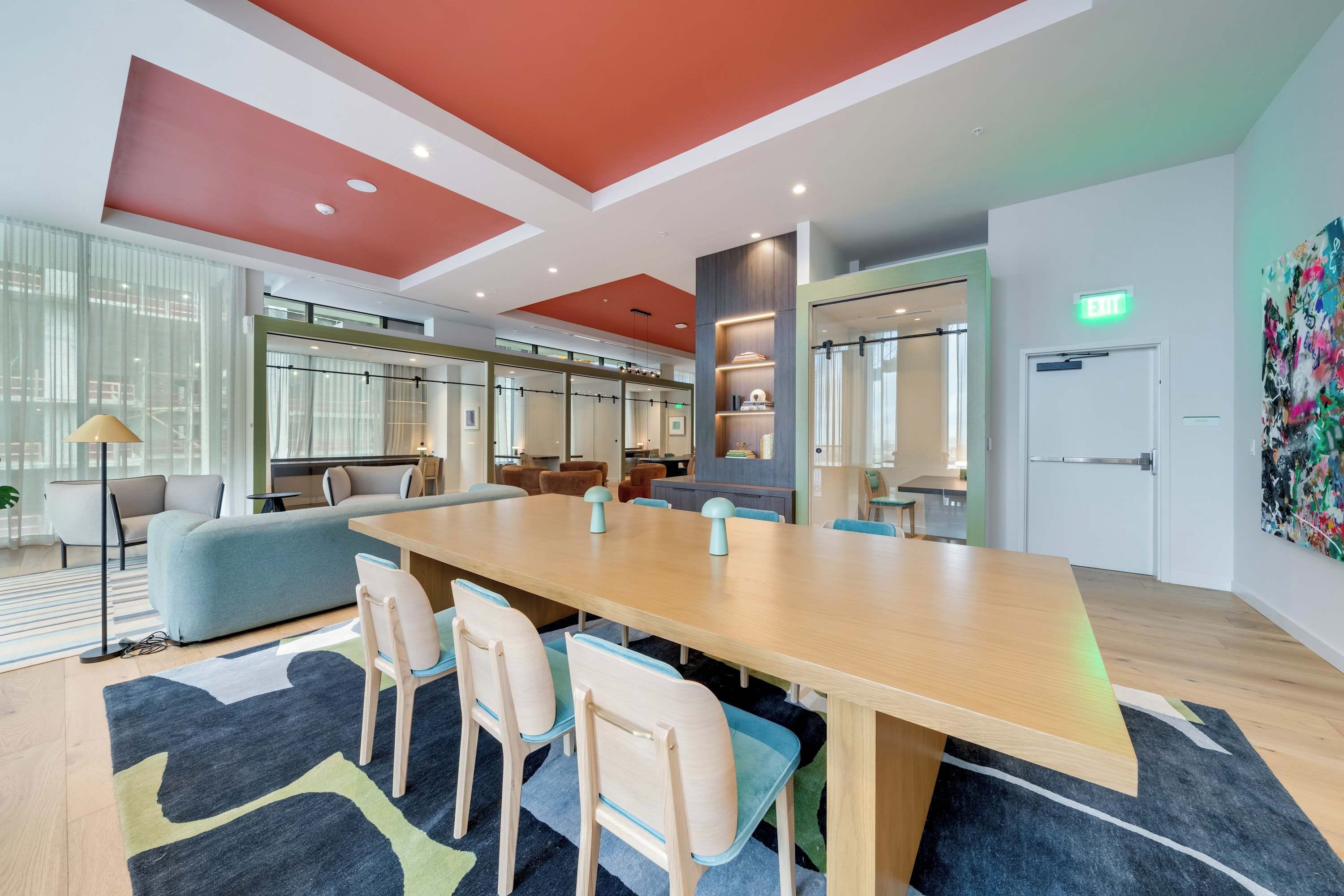A modern conference room with a long table, chairs, and a sofa. There are large windows, a ceiling with recessed lighting, and a door with an exit sign.