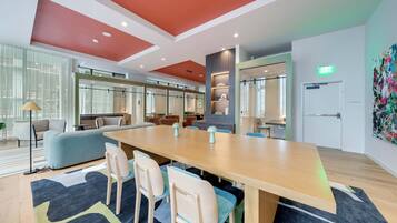 A modern conference room with a long table, chairs, and a sofa. There are large windows, a ceiling with recessed lighting, and a door with an exit sign.