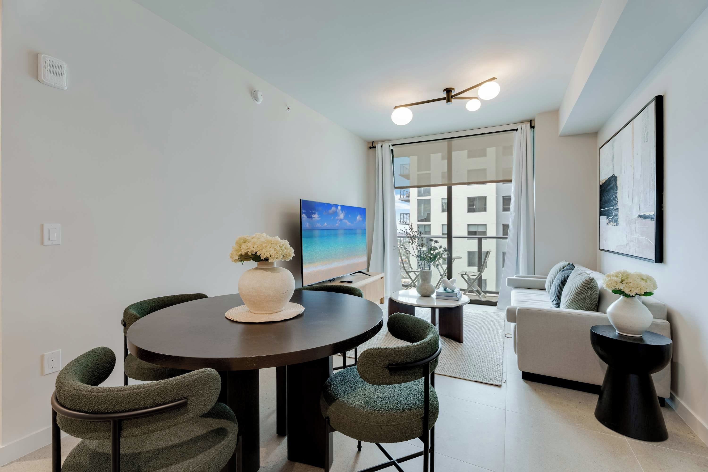 Una sala de estar moderna con una mesa de comedor redonda, sillas tapizadas en verde, un sofá y un ventanal con vistas a edificios.