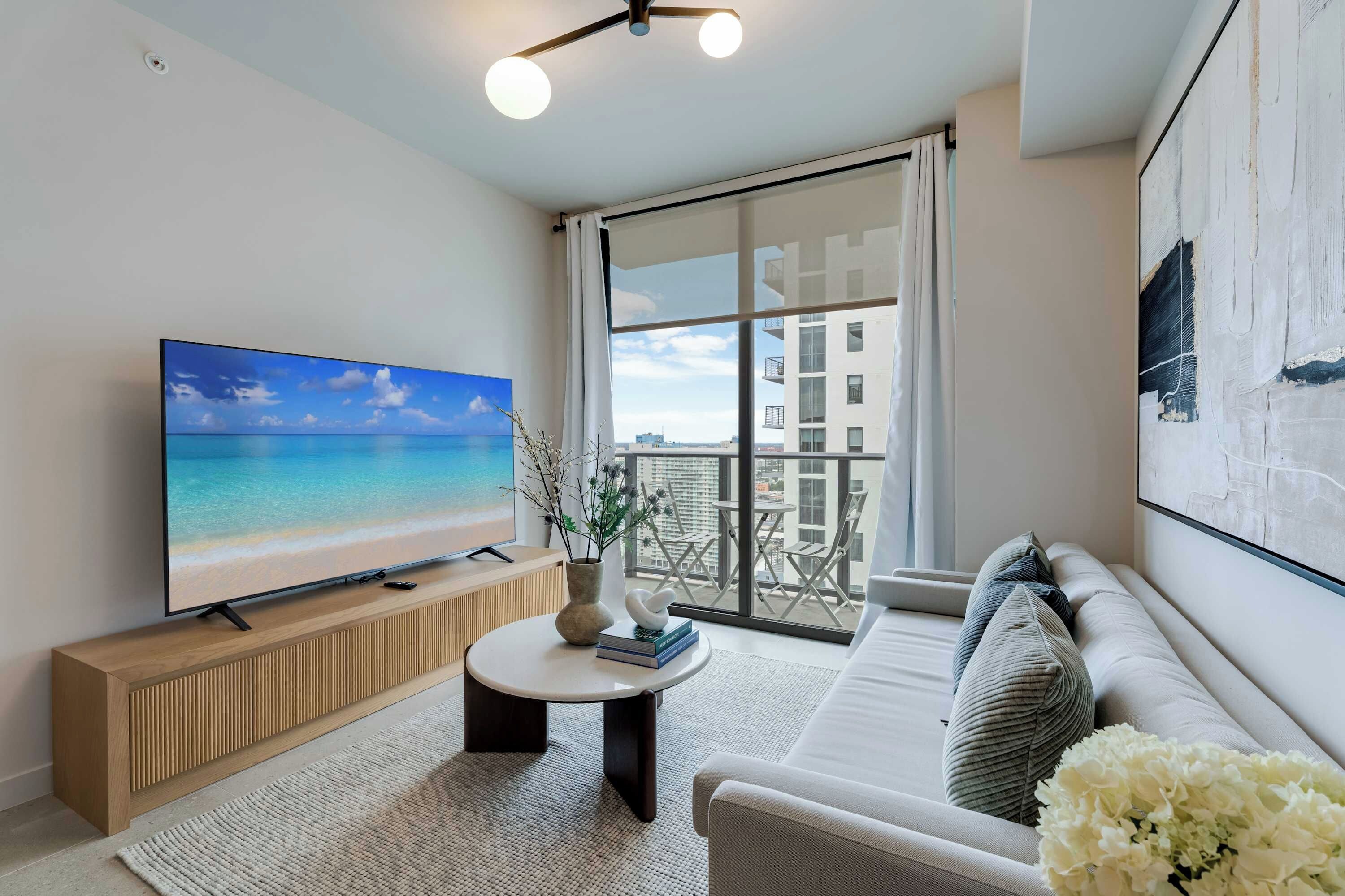 A modern living room with a large flat-screen TV, a wooden console table, a round coffee table, a sofa, and a large window with a balcony view.