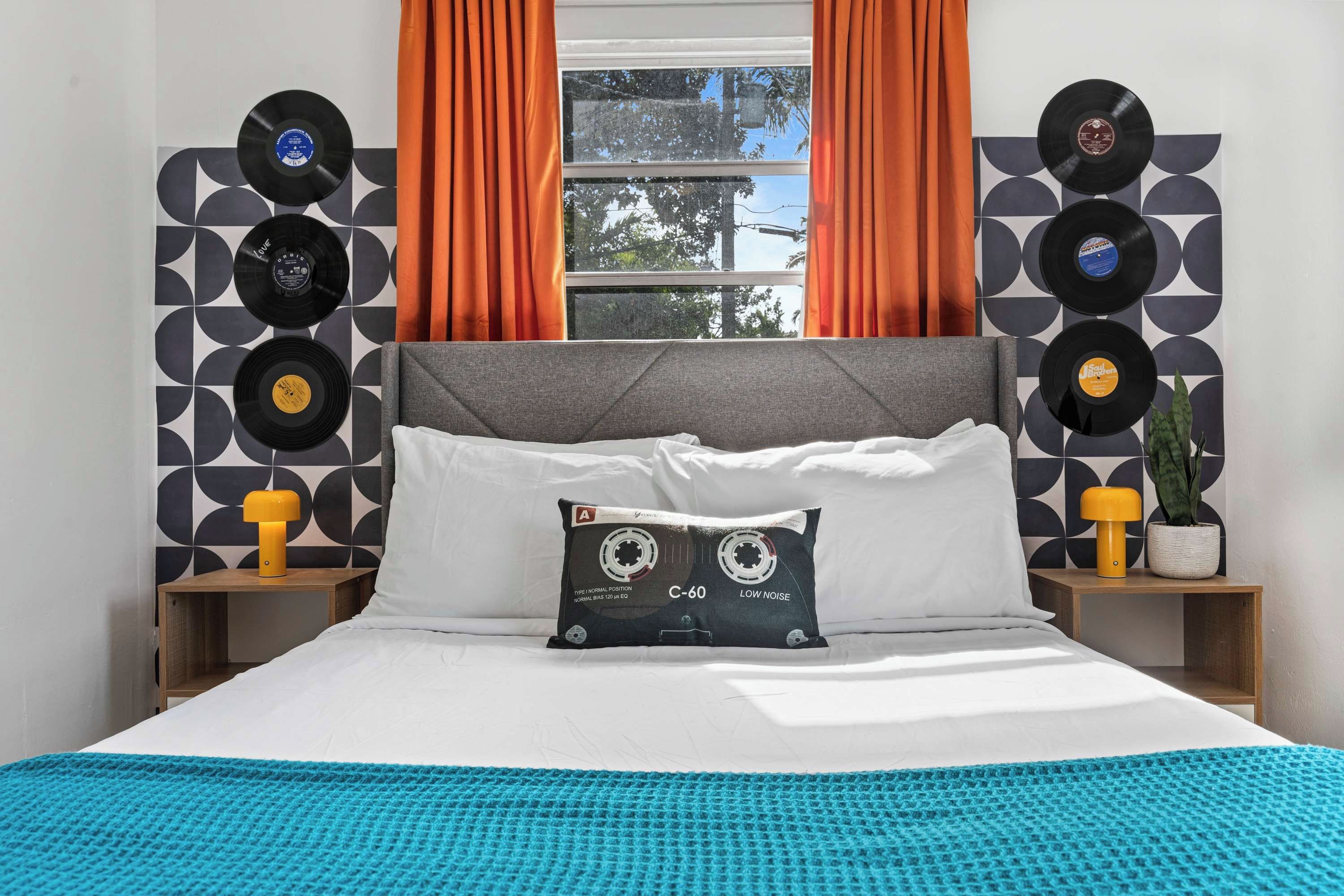 Uma cama com cabeceira cinzenta, almofadas brancas, manta azul e uma almofada com estampa de disco de vinil. Há duas mesas de cabeceira com luminárias e um vaso de planta.